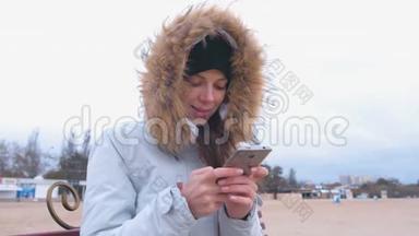 穿着白色<strong>羽绒服</strong>的女人坐在海滩上的长凳上，在手机上输入一条信息。